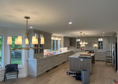 Spacious modern kitchen with light wood floors, white cabinets, large island, golden chandeliers, and various cleaning supplies on countertops and floor.