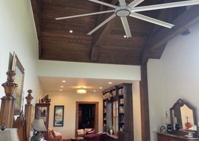 A bedroom with a high wooden ceiling, a large ceiling fan, a bed with pillows, a dresser with a mirror, and a sitting area with bookshelves.