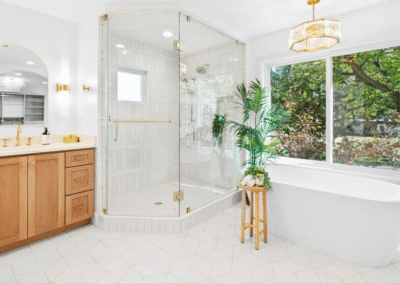 Bright bathroom with a glass shower, wooden vanity, freestanding tub, large windows, potted plant, and gold fixtures.