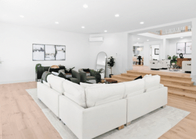 Modern living room with a white sectional sofa, wooden floors, indoor plants, and wall art. Room features open layout with steps leading to the dining area in the background.