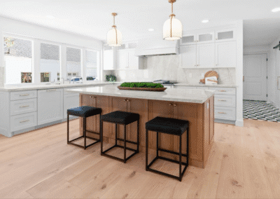 Modern kitchen with a large island, three black cushioned stools, white cabinets, pendant lights, and wood flooring. Marble backsplash and countertops are also visible.