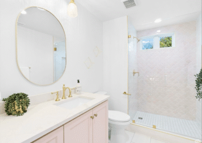 Modern bathroom with a round mirror, single sink vanity, gold fixtures, glass-enclosed shower, and light pink cabinetry.