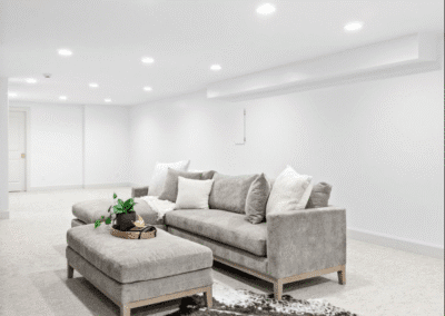 A modern basement living area with white walls, ceiling lights, a gray sofa, a matching ottoman, and a decorative tray with a plant on top.