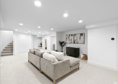 Modern basement living area with light gray sectional sofa, wall-mounted artwork, media console, and carpeted floors under recessed lighting. Stairs visible in the background.