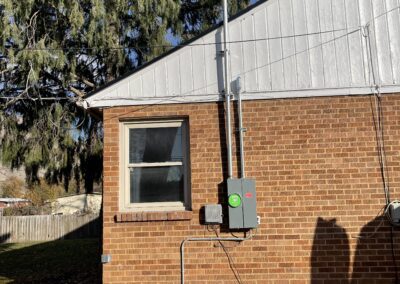 An electrical meter and service panel with conduit are mounted on the brick exterior wall of a house next to a window and beneath a sloped roof.