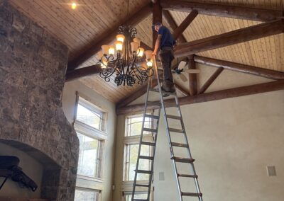 A man on a tall ladder adjusts a chandelier in a living room with high vaulted ceilings, large windows, and wooden beams.